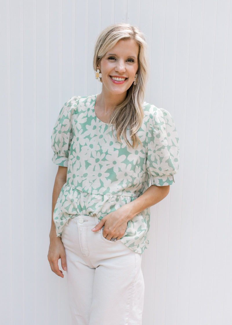 Model wearing daisy earrings with a sage babydoll top with cream daisies, with puff short sleeves.