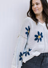 Close up of blue flowers with yellow center on an ivory long sleeve sweater with exposed hem. 