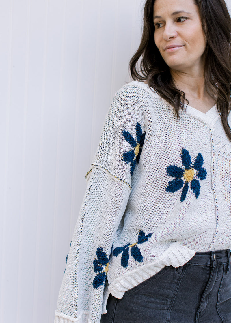 Close up of blue flowers with yellow center on an ivory long sleeve sweater with exposed hem. 