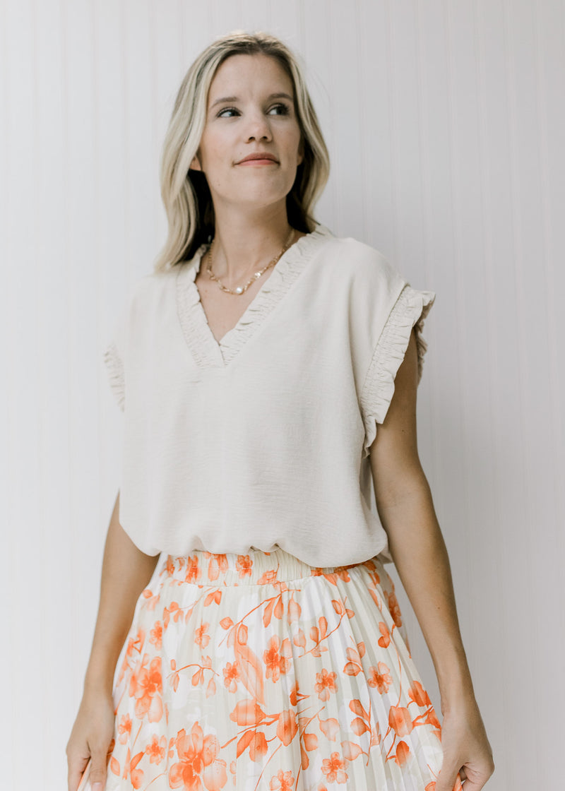 Model wearing an oatmeal top with a smocked detail at the v-neck and short sleeves tucked in a skirt