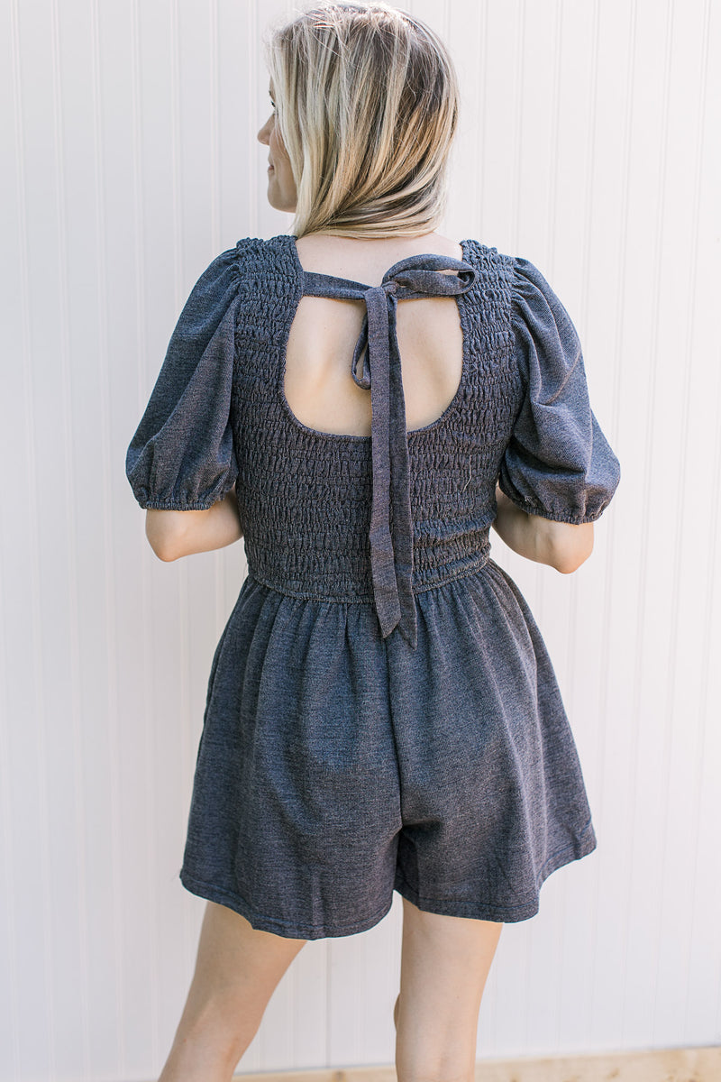 Model wearing a dark denim romper with a tie back, short sleeves and pleated front.