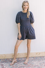 Model wearing sandals with a dark denim romper with pockets, short sleeves and pleated front.