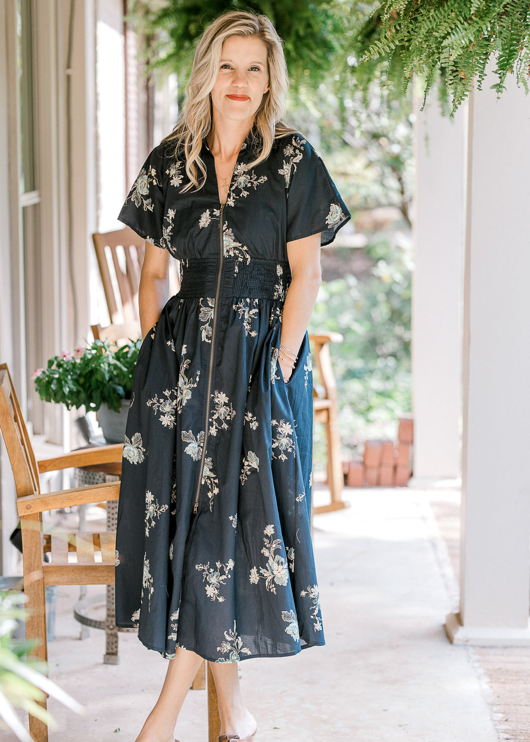 Model wearing a short sleeve black midi with cream flowers, pockets, smocked waist and v-neck. 