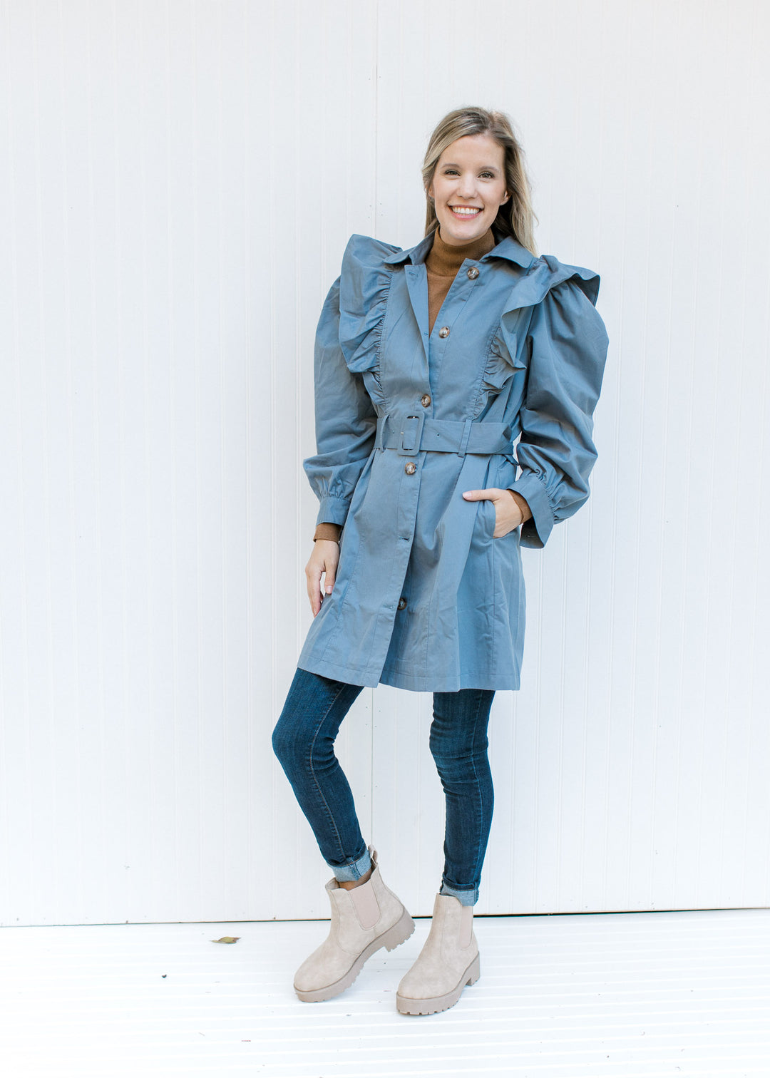 Model wearing jeans and booties with a vintage blue coat with a ruffle at the shoulder and a belt. 
