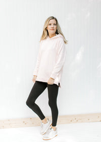 Model wearing black leggings and sneakers with a blush colored hoodie with long sleeves. 