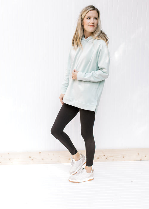Model wearing black leggings and sneakers with a mint hoodie with long sleeves and a side zippers. 