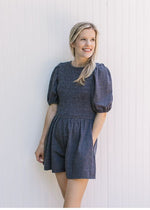 Model wearing a dark denim romper with pockets, short sleeves and pleated front.