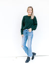 Model wearing jeans and booties with a hunter green cable knit sweater with and long sleeves. 