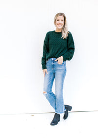 Model wearing jeans and booties with a hunter green cable knit sweater with and long sleeves. 