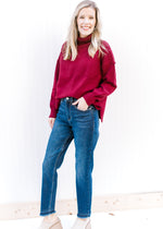 Model wearing jeans, mules and a deep red sweater with a loose turtle neck and long sleeves. 