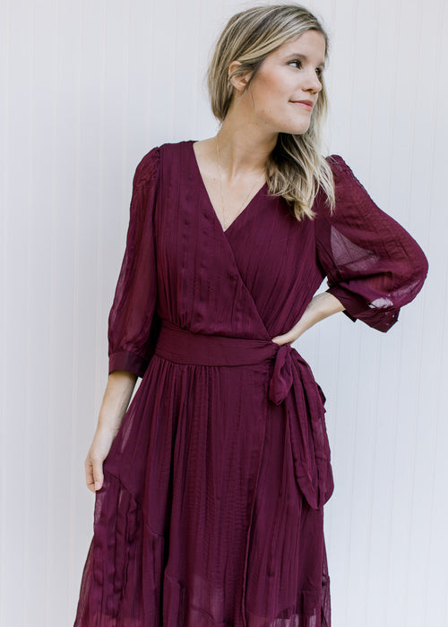 Model wearing a wine colored wrap dress with a v-neck, sheer 3/4 sleeves and a pleated material. 
