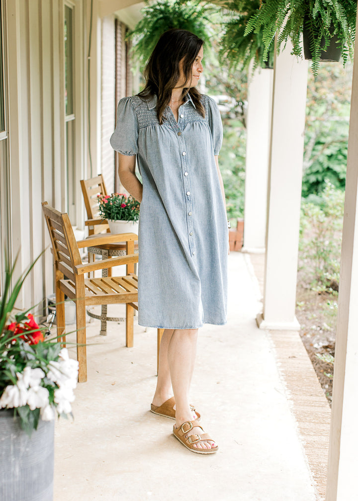 Model wearing a light wash denim dress with bubble short sleeves, smocked yoke and a button front.