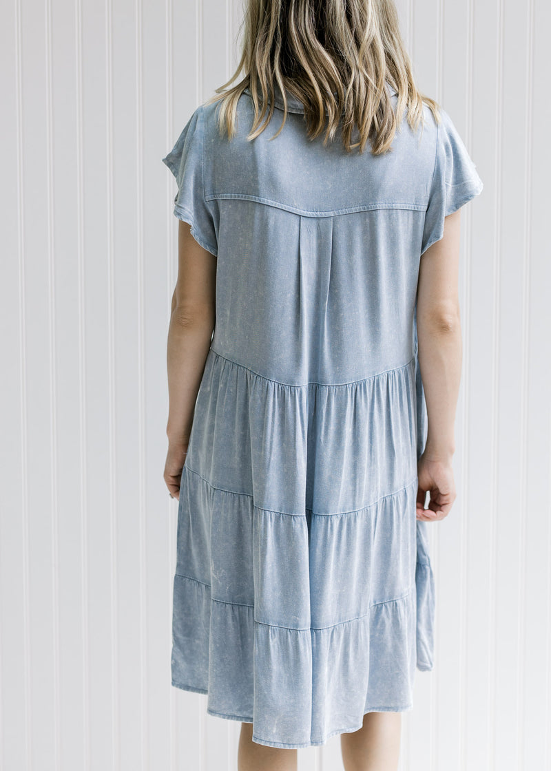 Back view of Model wearing a light denim button up dress with short sleeves, hitting above the knee
