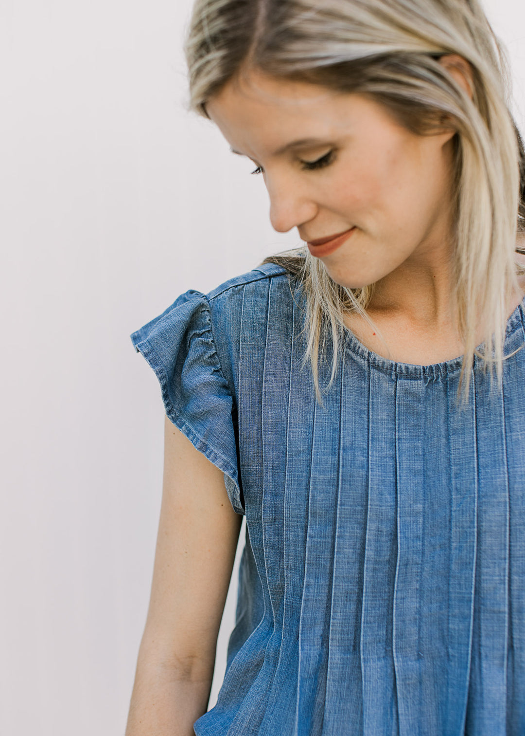 Model wearing a medium/dark denim top with flutter cap sleeves, a pleated design and round neck.