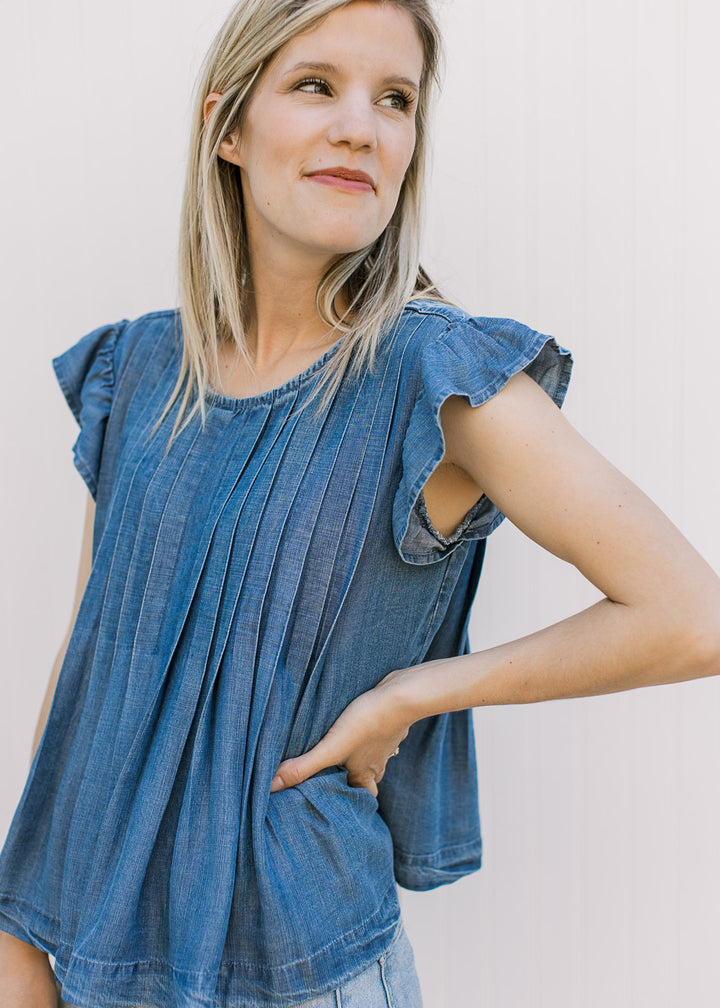 Model wearing a flowy medium/dark denim top with flutter cap sleeves, and pleated design.