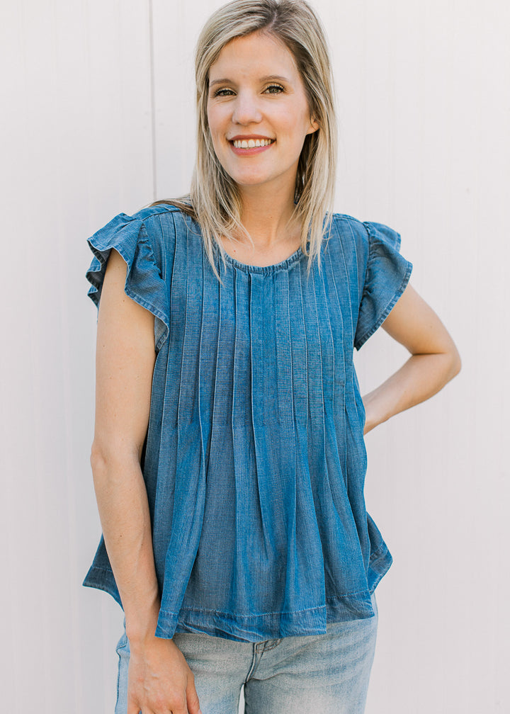 Model wearing jeans and a medium/dark denim top with flutter cap sleeves, pleating and round neck.