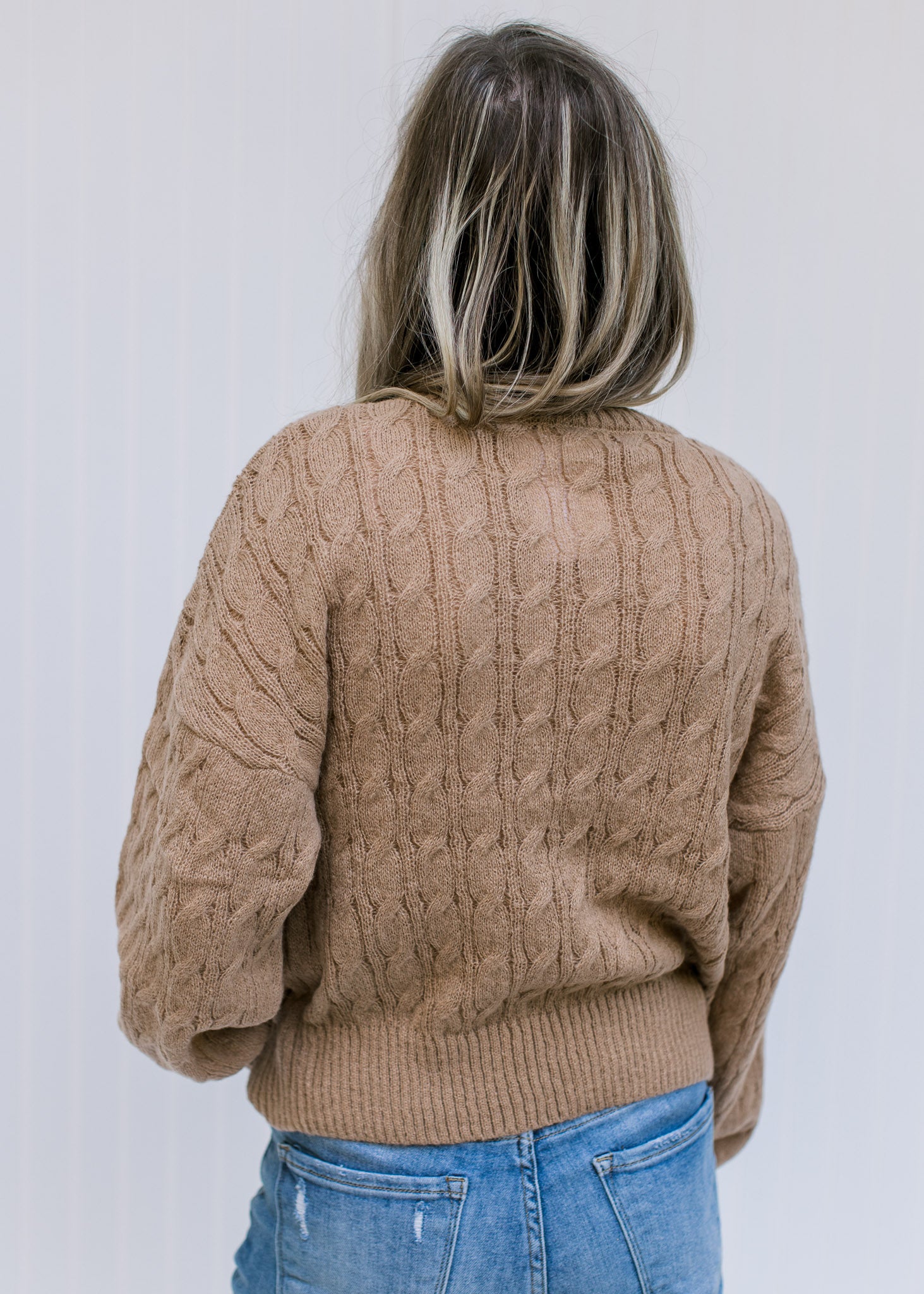 Back view of Model wearing a camel sweater with a round neck, long sleeves and a cable knit fabric.