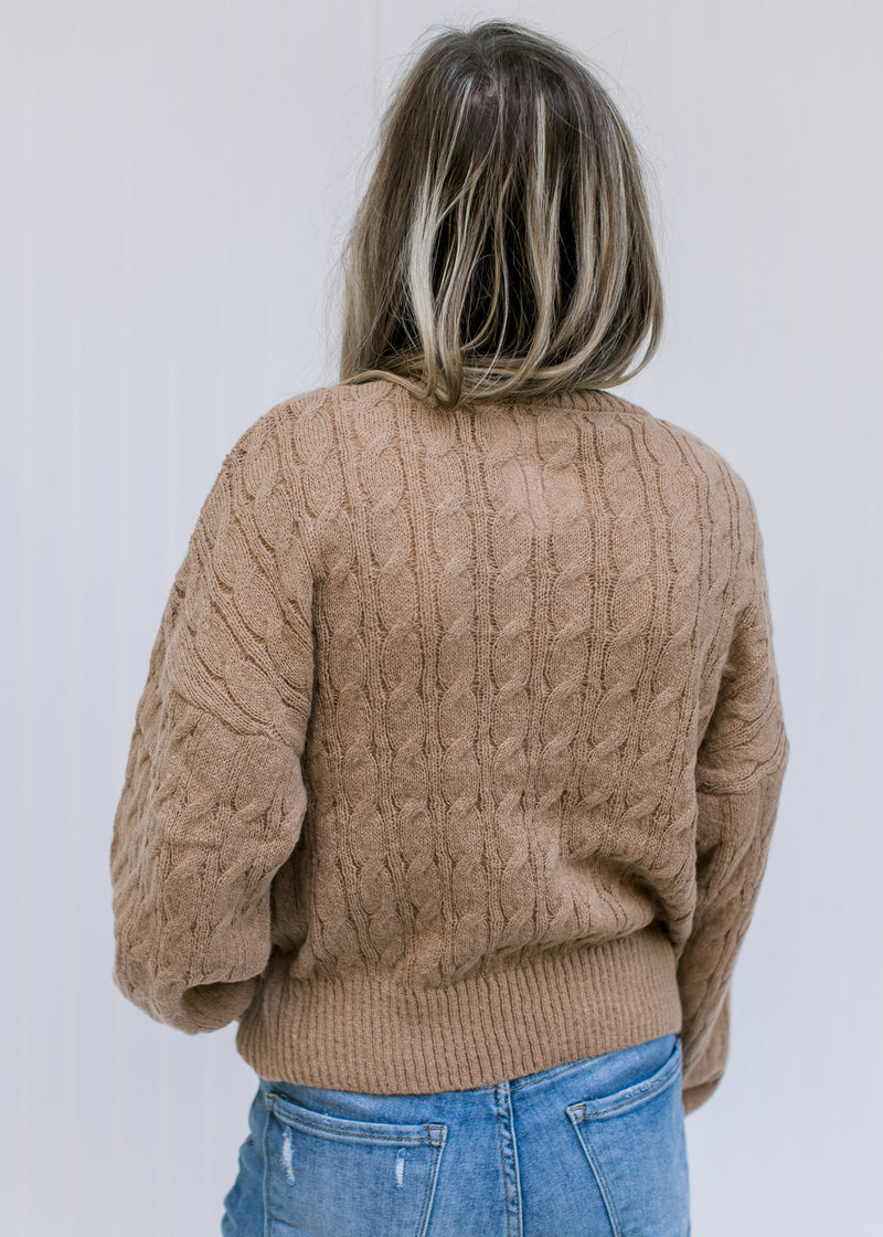 Back view of Model wearing a camel sweater with a round neck, long sleeves and a cable knit fabric.
