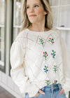 Model wearing a cream diamond open work sweater with embroidered flower motif.