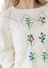 Close up of embroidered flower motif on a cream long sleeve sweater with diamond pattern. 