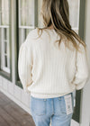 Back view of Model wearing a long sleeve cream sweater with embroidered flower motif.