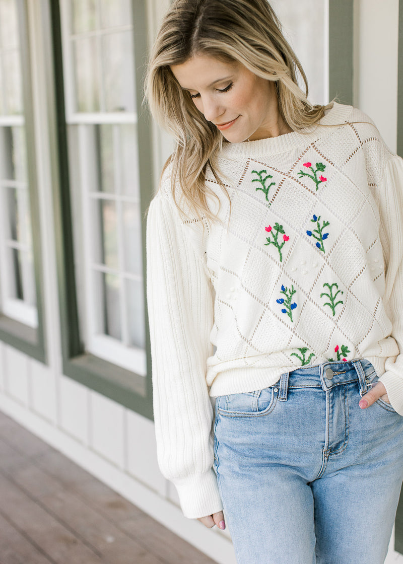 Model wearing jeans and a cream diamond open work sweater with embroidered flower.