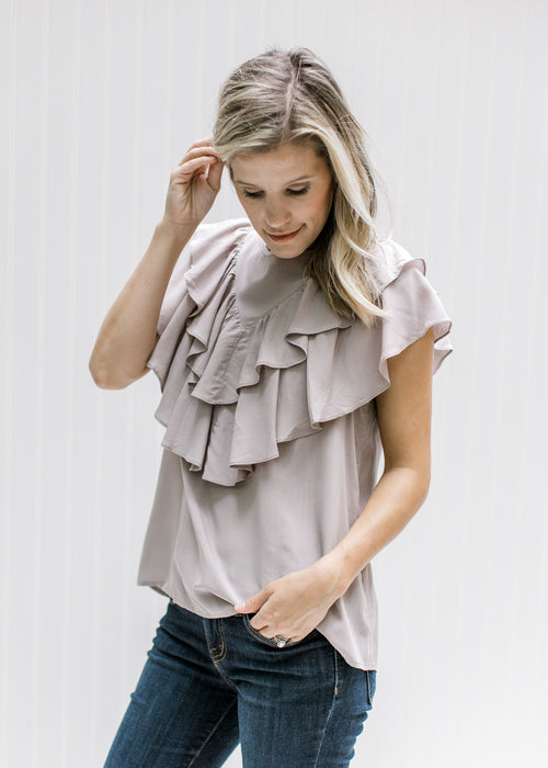 Model wearing a stone colored top with a button up back, round neck and double ruffle detail. 