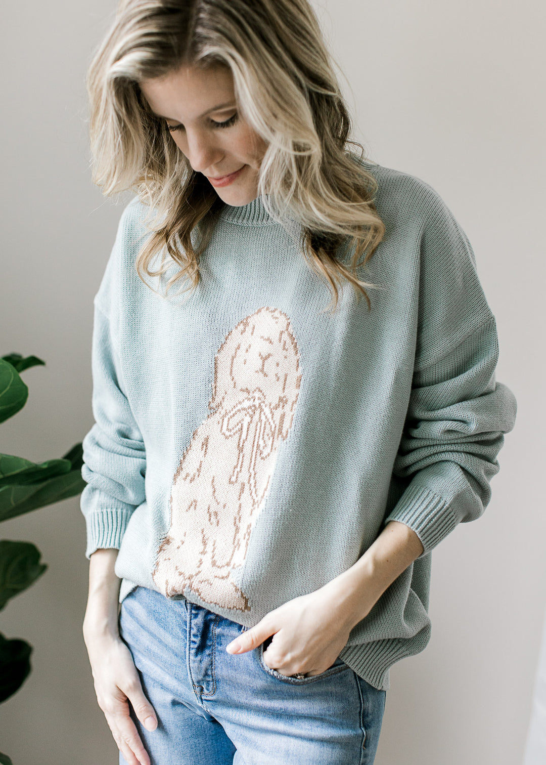 Model wearing a long sleeve blue-green sweater with bunny detail on the front and round neck. 