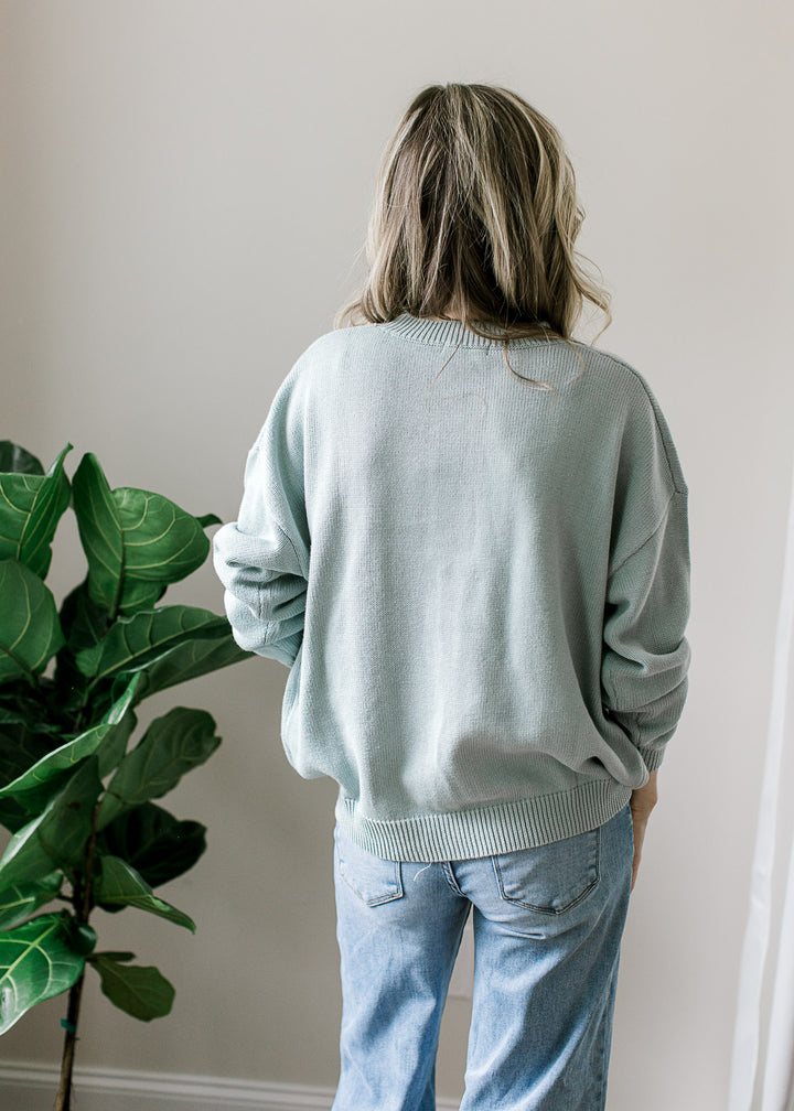 Back view of Model wearing a long sleeve blue-green sweater with bunny detail on the front. 