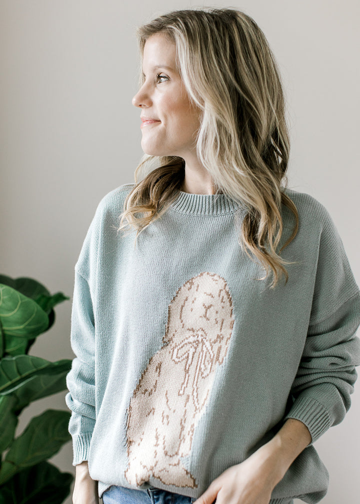 Model wearing a long sleeve blue-green sweater with taupe bunny detail on the front and round neck. 
