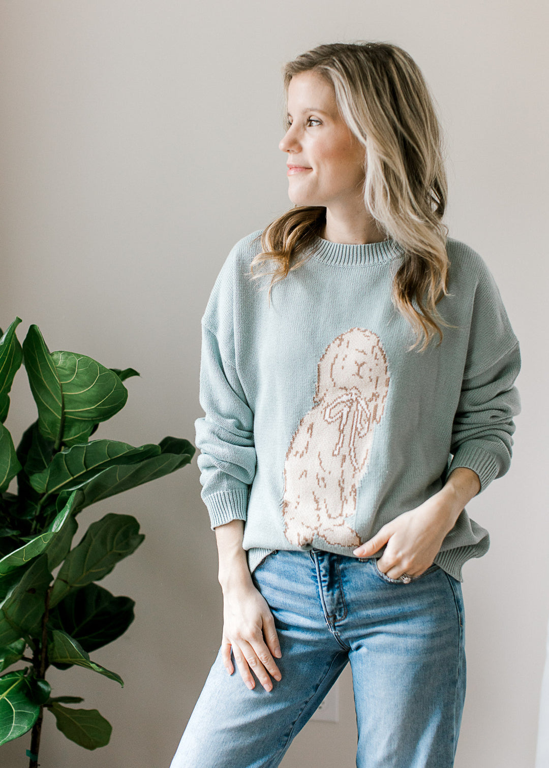 Model wearing jeans and a long sleeve blue-green sweater with bunny detail on the front. 