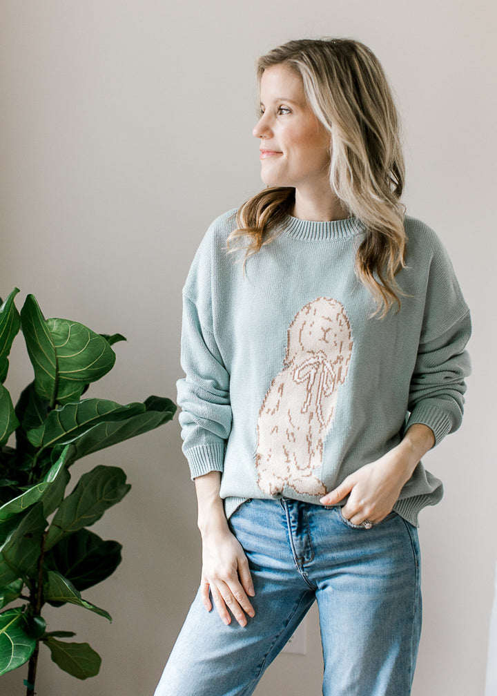 Model wearing jeans and a long sleeve blue-green sweater with bunny detail on the front. 