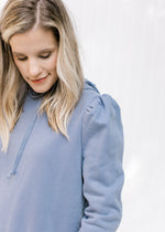 Close up view of puff shoulders on a model in a dusty blue pullover with long sleeves and a hoodie. 