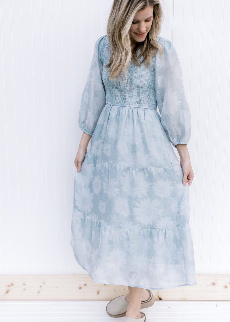 Model wearing a sage v-neck dress with a smocked bodice, 3/4 sheer sleeves and a faint daisy print.