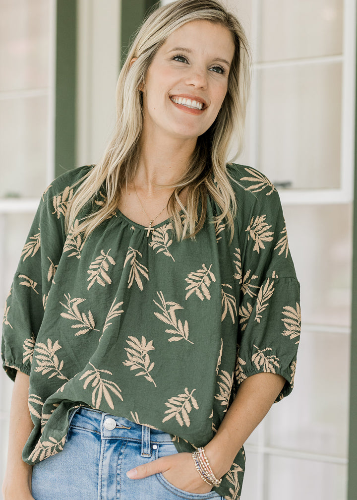 Model wearing a flowy hunter green top with wheat colored graphics, v-neck and short sleeves.