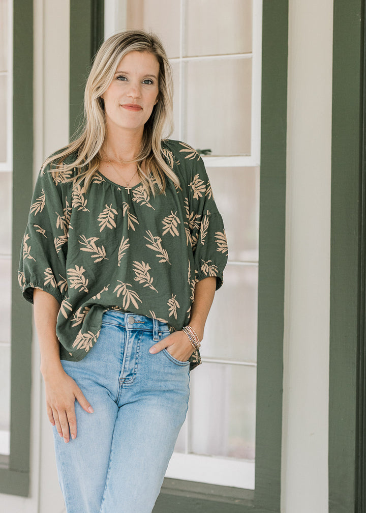 Model wearing jeans and a hunter green top with wheat colored graphics, v-neck and short sleeves.