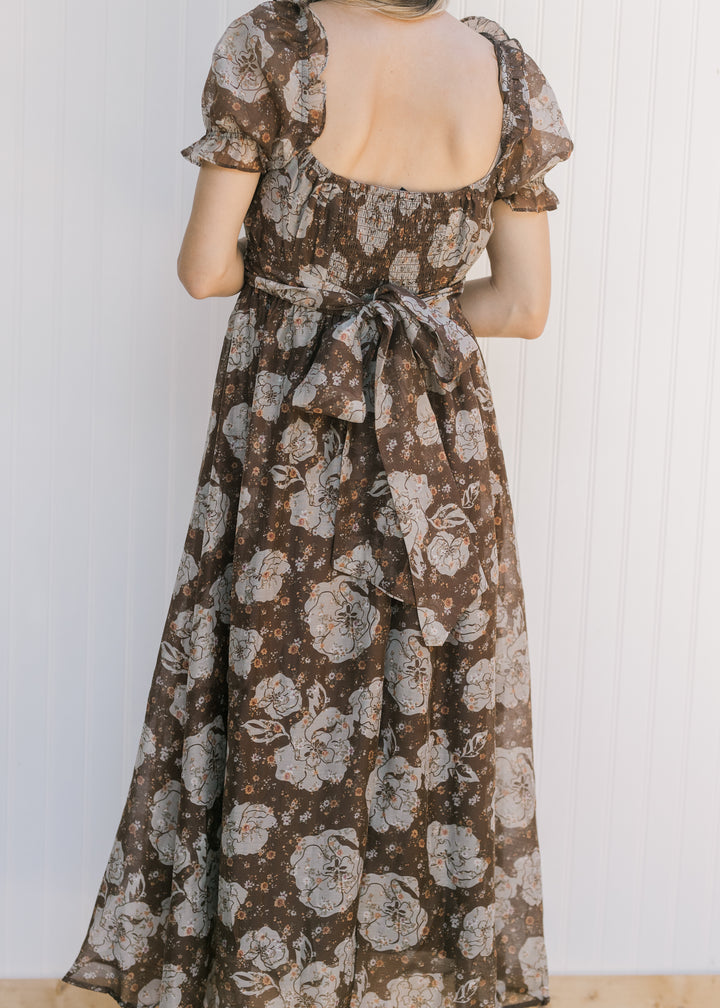 Back view of a model wearing a brown midi with floral, square neck and smocked back with tie. 