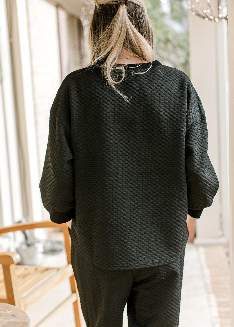 Back view of model in a black textured top with 3/4 sleeves and round neck with matching pants.