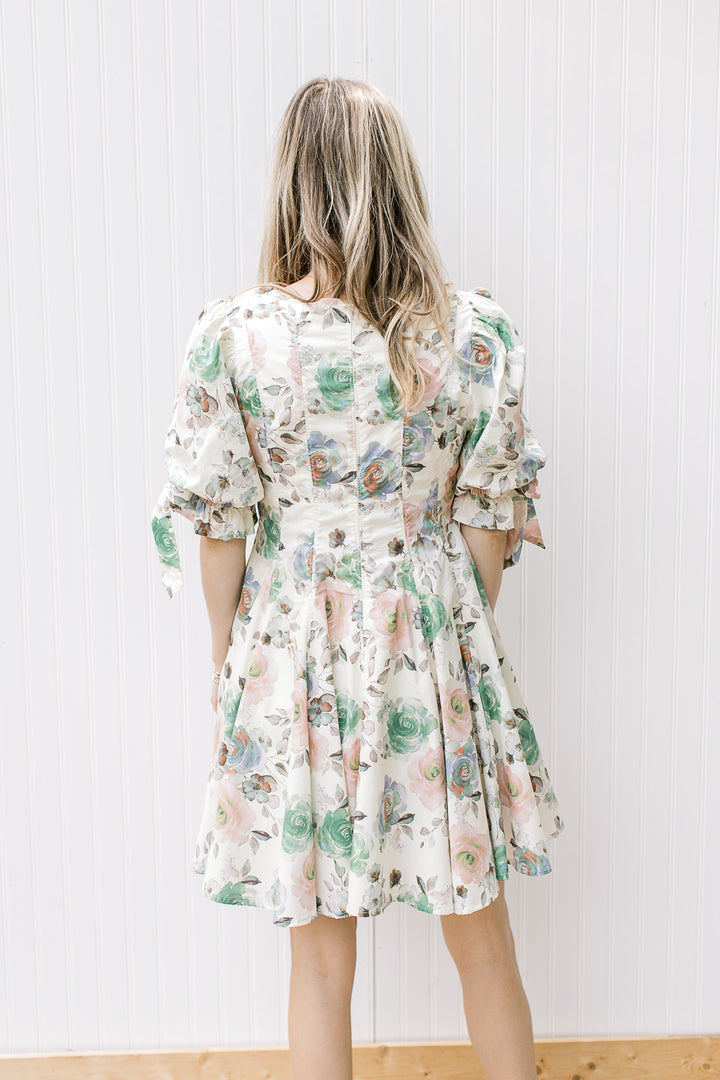 Back view of Model wearing a short sleeve cream dress with watercolor floral and pleated bodice. 