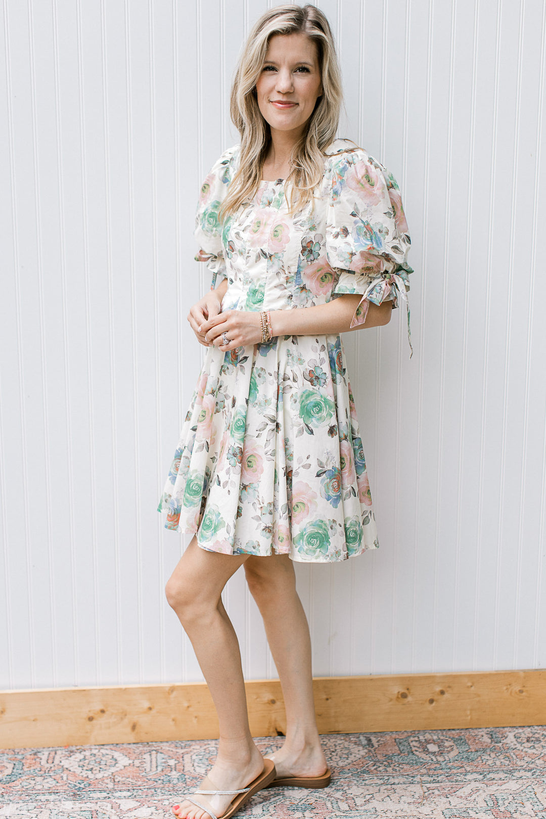 Model wearing sandals and a cream above the knee dress with watercolor floral and pleated bodice. 