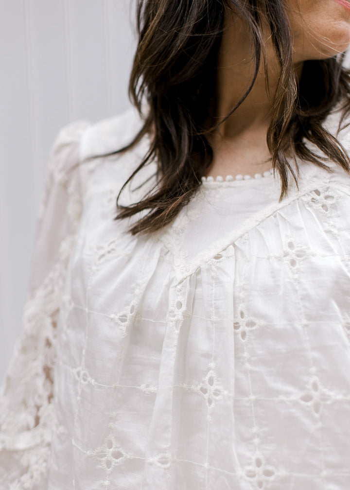 Model wearing a cream top with embroidered flowers, eyelet and sheer long sleeves. 