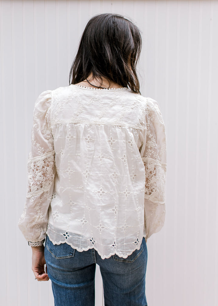 Back view of Model wearing a cream top with embroidered flowers, eyelet and sheer long sleeves. 