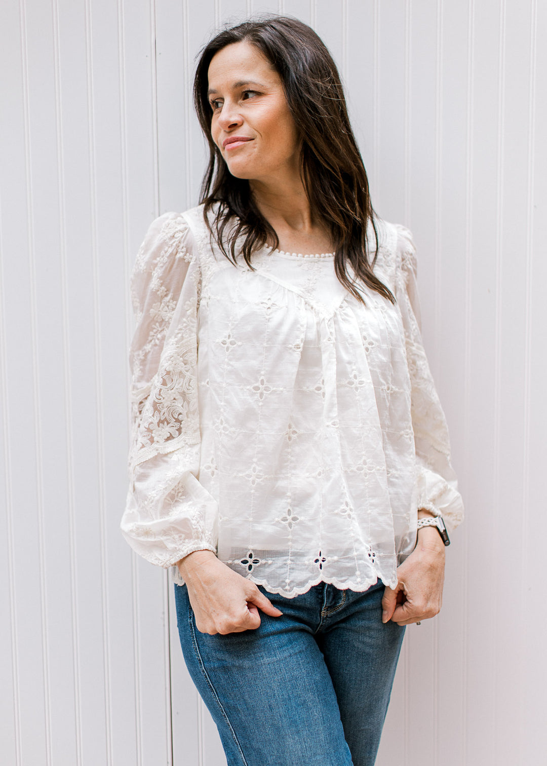 Model wearing jeans and a cream top with embroidered flowers, eyelet and sheer long sleeves. 