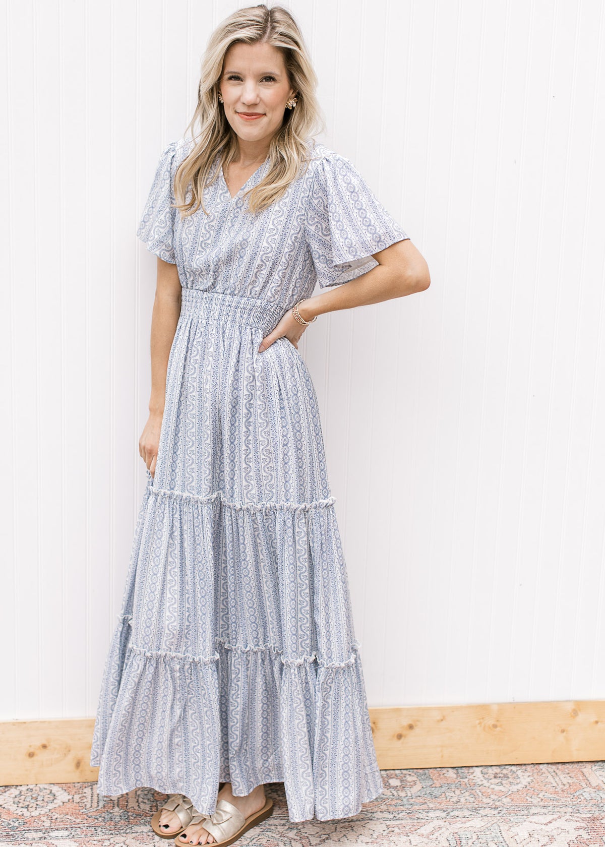Model wearing sandals and a tiered white maxi with a pale blue print and a wide elastic waist. 