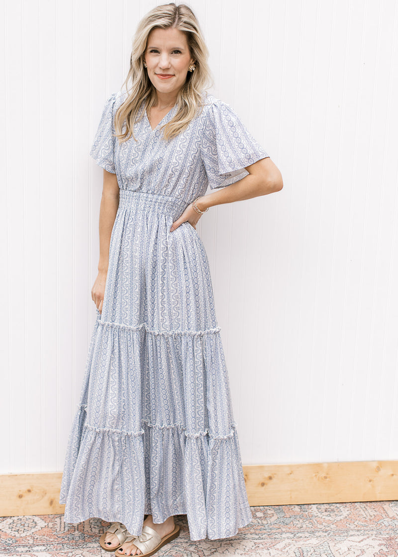 Model wearing sandals and a tiered white maxi with a pale blue print and a wide elastic waist. 