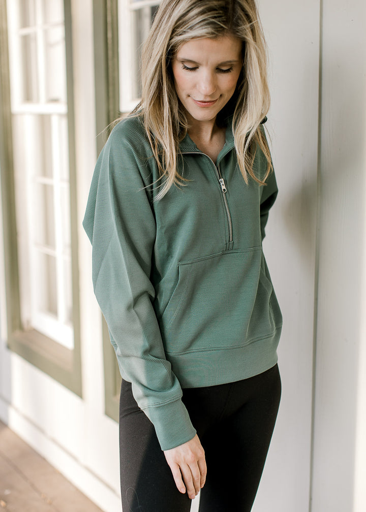Model wearing a dark green ribbed pullover with a quarter zip, front pouch pocket and long sleeves. 