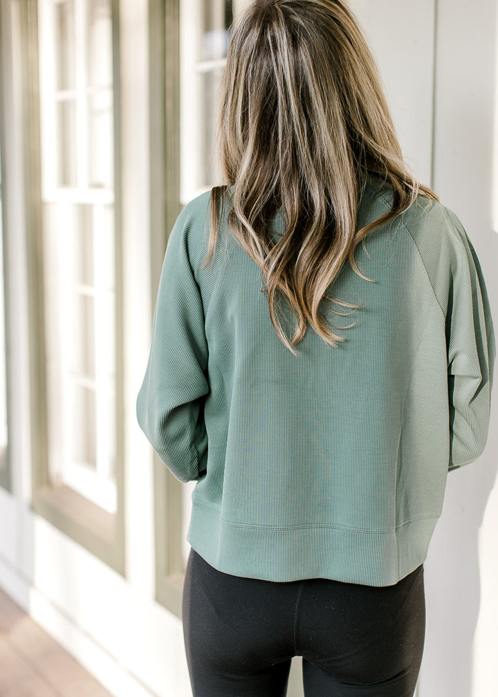 Back view of Model wearing a dark green ribbed pullover with a front pouch pocket and long sleeves. 