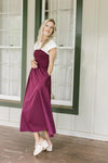 Model wearing wedges and a plum dress with a smocked bodice, white yolk with cap short sleeves. 