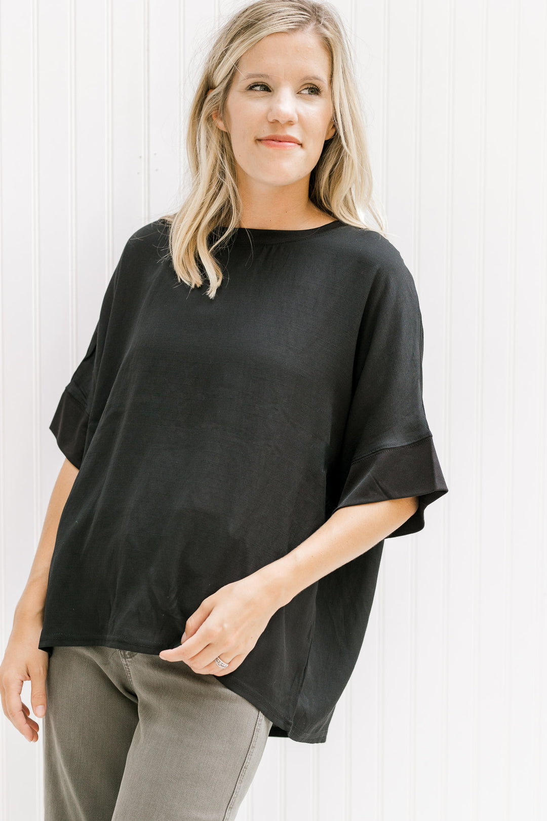 Model wearing gray pants with a black top with a round neck and short sleeves with a ruffle detail.