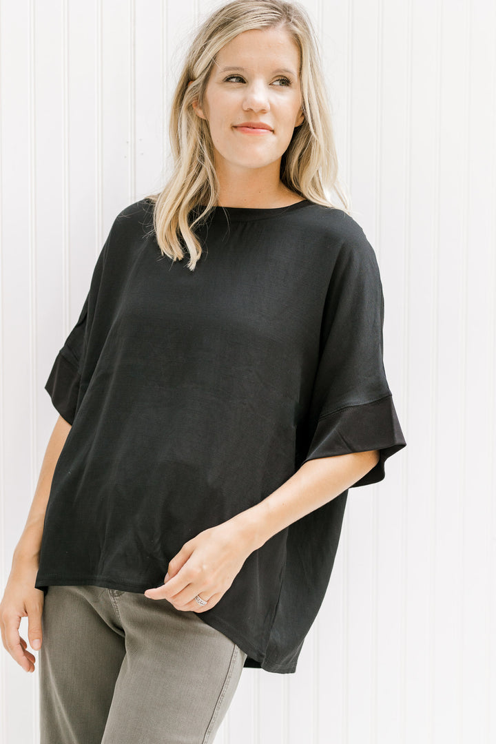 Model wearing gray pants with a black top with a round neck and short sleeves with a ruffle detail.